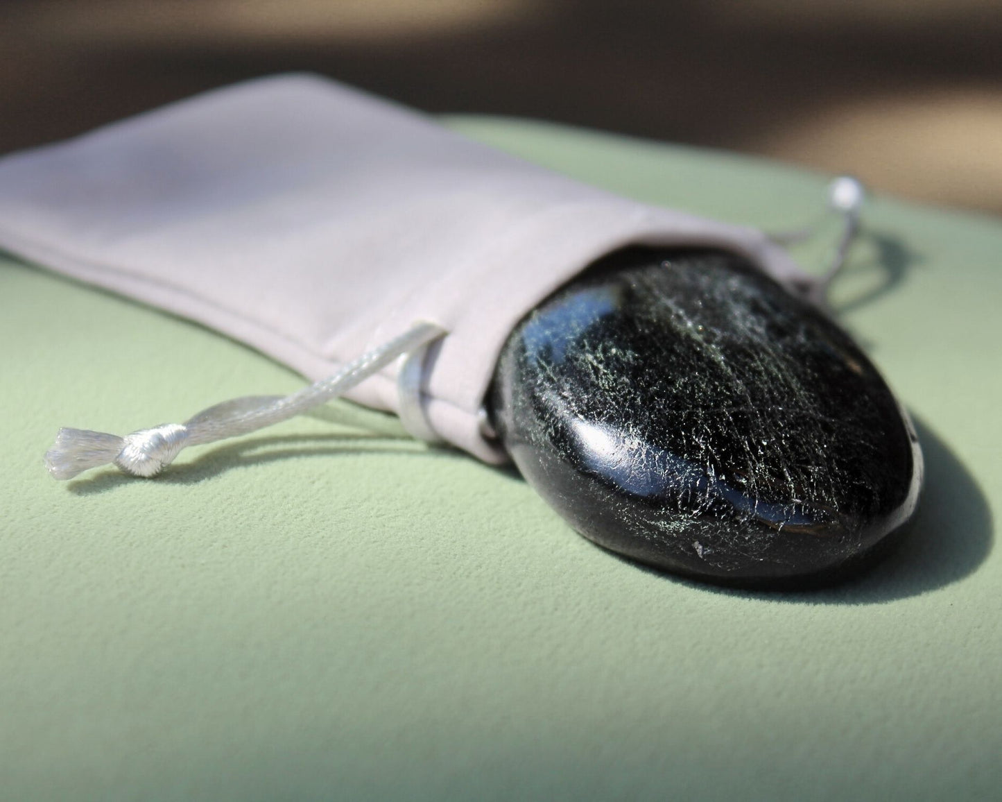 Black Tourmaline palm stone partially out of a grey velvet drawstring bag, displayed on a sage green cushion — used for meditation, grounding, and energy protection.
