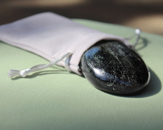 Black Tourmaline palm stone partially out of a grey velvet drawstring bag, displayed on a sage green cushion — used for meditation, grounding, and energy protection.