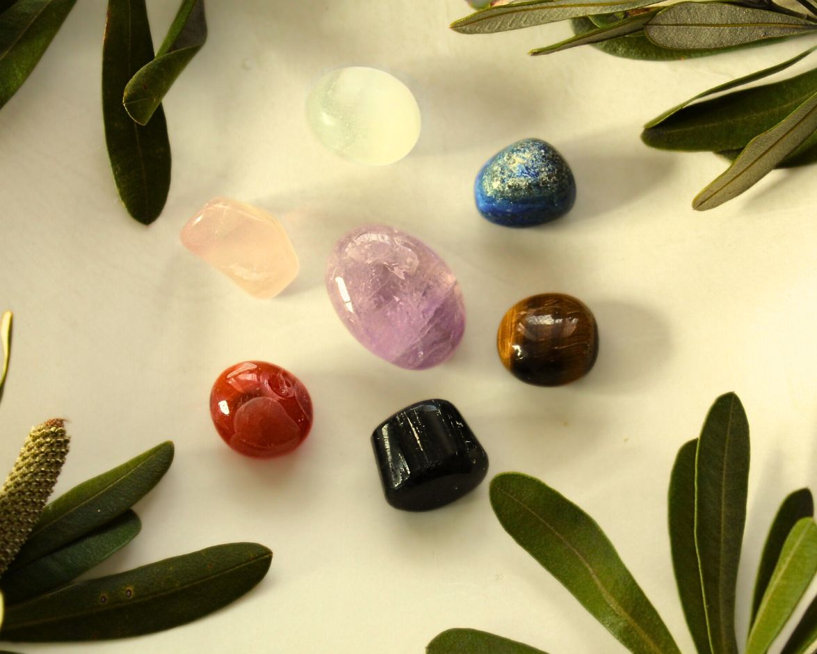 Seven tumbled chakra crystals from the 7 Chakra Meditation Kit displayed on a light yellow background with natural greenery — each crystal represents a chakra and is used for meditation, energy balancing, and mindfulness practices.