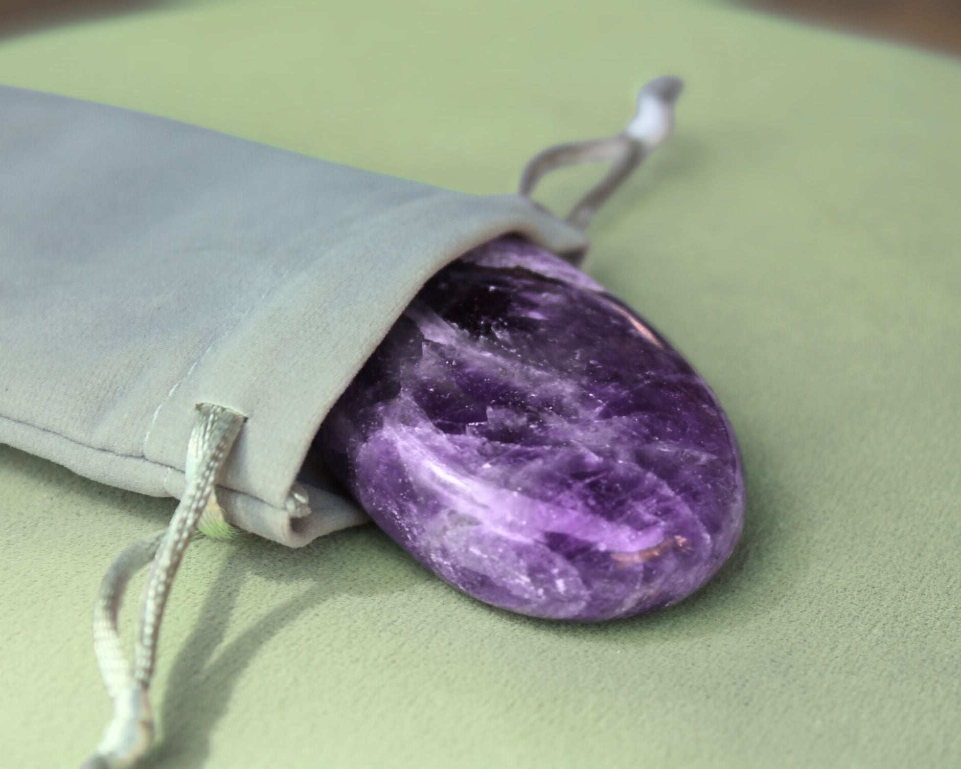 Amethyst palm stone partially out of a grey velvet drawstring bag, displayed on a sage green cushion — used for meditation, energy healing, and promoting calm, intuition, and spiritual awareness.