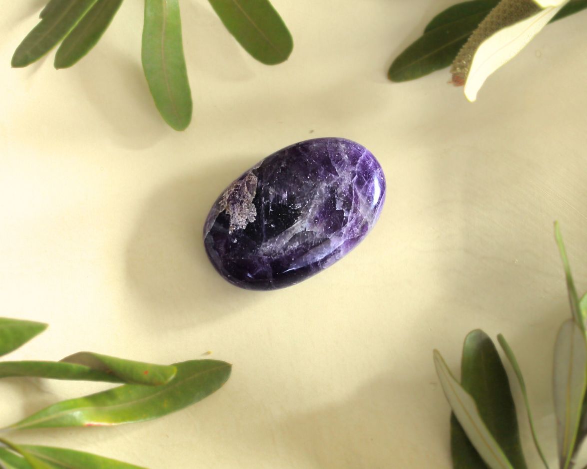 Amethyst palm stone displayed on a light yellow background with natural greenery — used for meditation, energy healing, and promoting calm, intuition, and spiritual awareness.