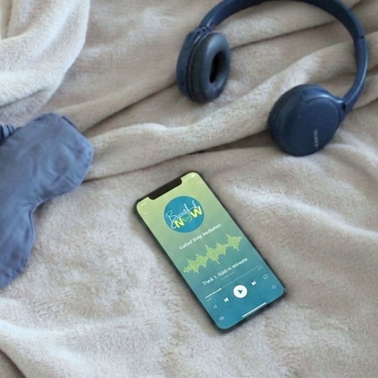 A smartphone displaying the Breath of NOW sleep meditation track on screen, placed alongside a blue eye mask and navy headphones on a soft, cream-coloured textured blanket.