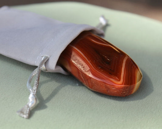 Carnelian palm stone partially out of a grey velvet drawstring bag, displayed on a sage green cushion — used for meditation, creativity, motivation, and vitality.