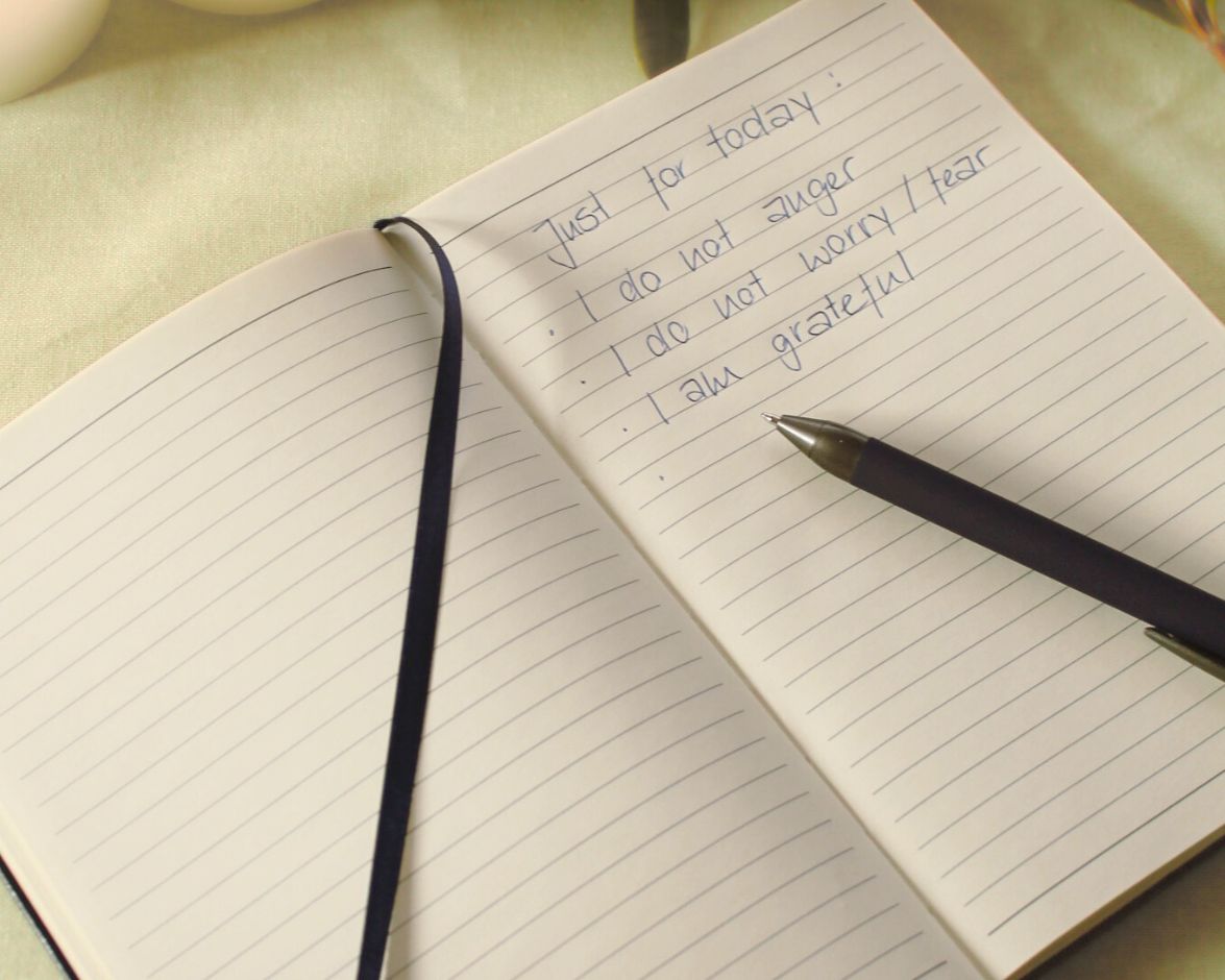Open Chakra Meditation Kit journal displayed on a light yellow background with natural greenery — pen resting on page with written affirmations: ‘Just for today – I do not anger, I do not worry/fear, I am grateful’ — for mindfulness, reflection, and intention setting as part of the 7 Chakra Meditation Kit.