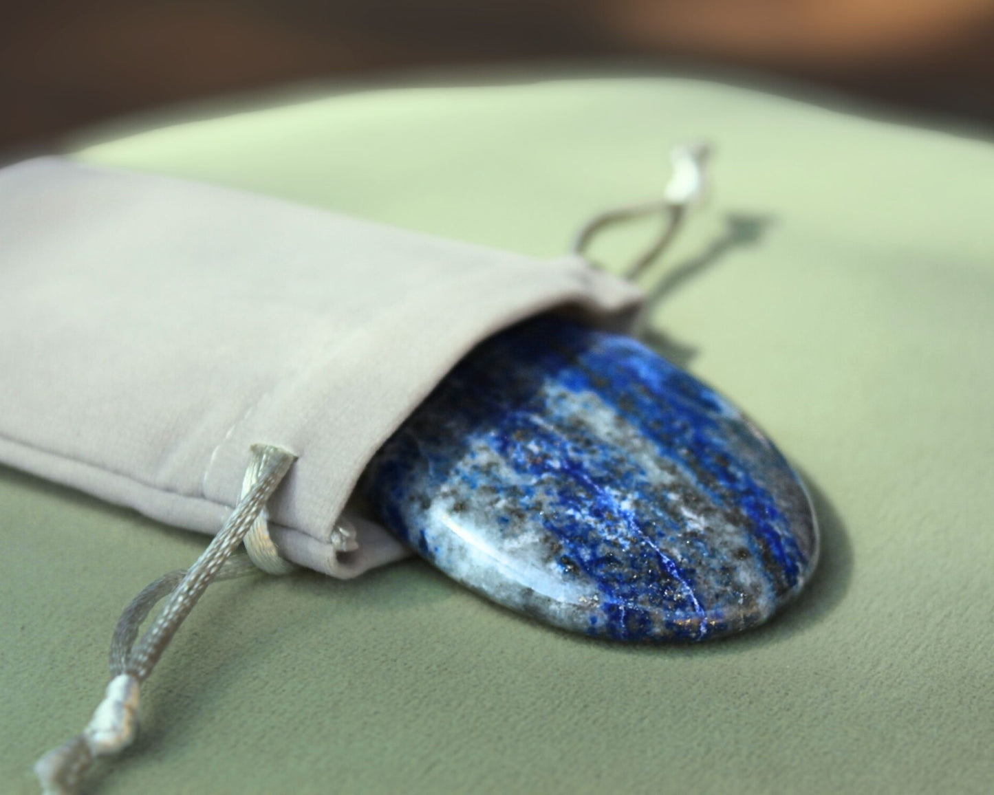 Lapis Lazuli palm stone partially out of a grey velvet drawstring bag, displayed on a sage green cushion — used for meditation, energy balancing, and promoting truth, self-expression, and clear communication.