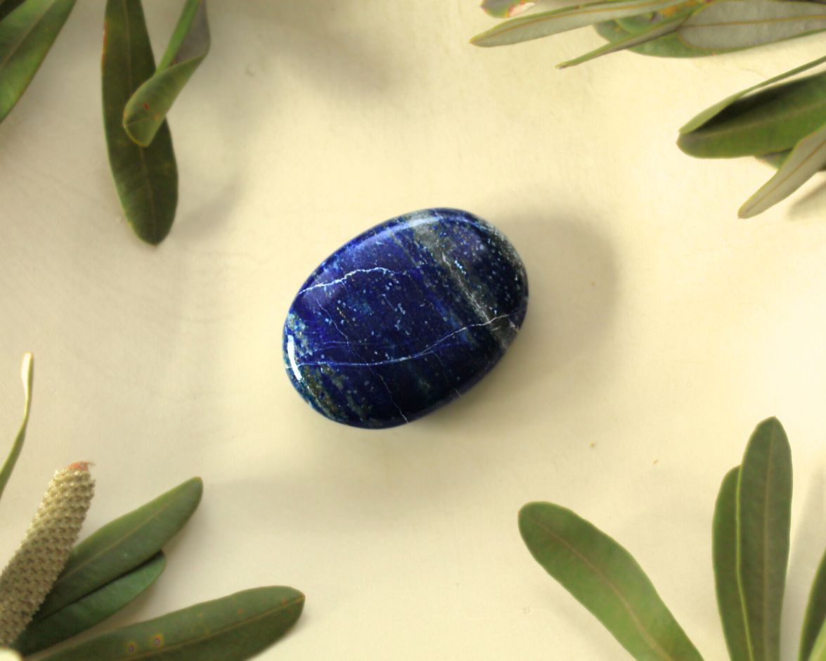 Lapis Lazuli palm stone displayed on a light yellow background with natural greenery — used for meditation, energy balancing, and promoting truth, self-expression, and clear communication.