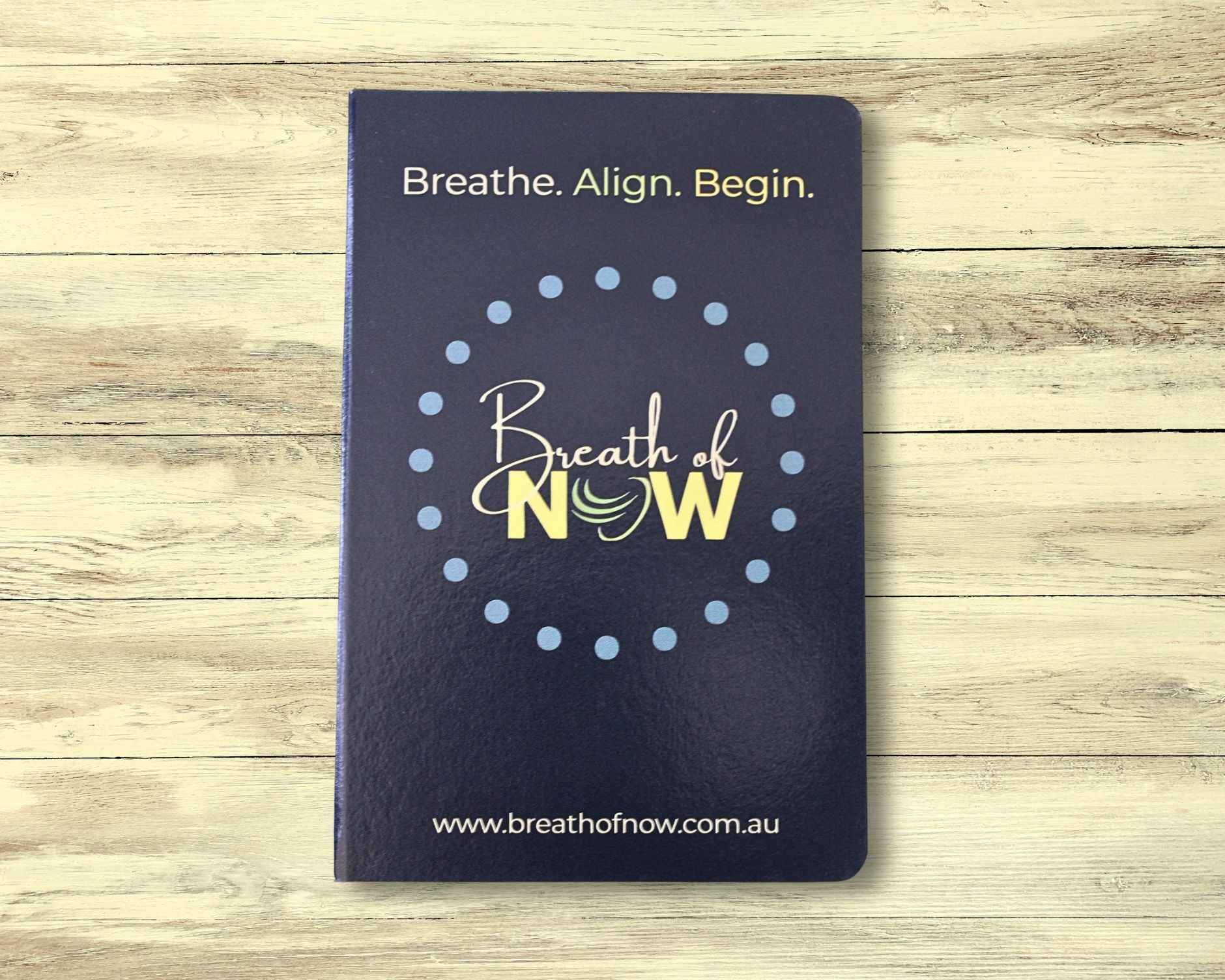 Front cover of dark blue meditation journal on light wooden floorboards — features text ‘Breath. Align. Begin.’, the Breath of NOW logo surrounded by a dotted circle, and the website ‘www.breathofnow.com.au’
 — designed for mindfulness, reflection, and intention setting.