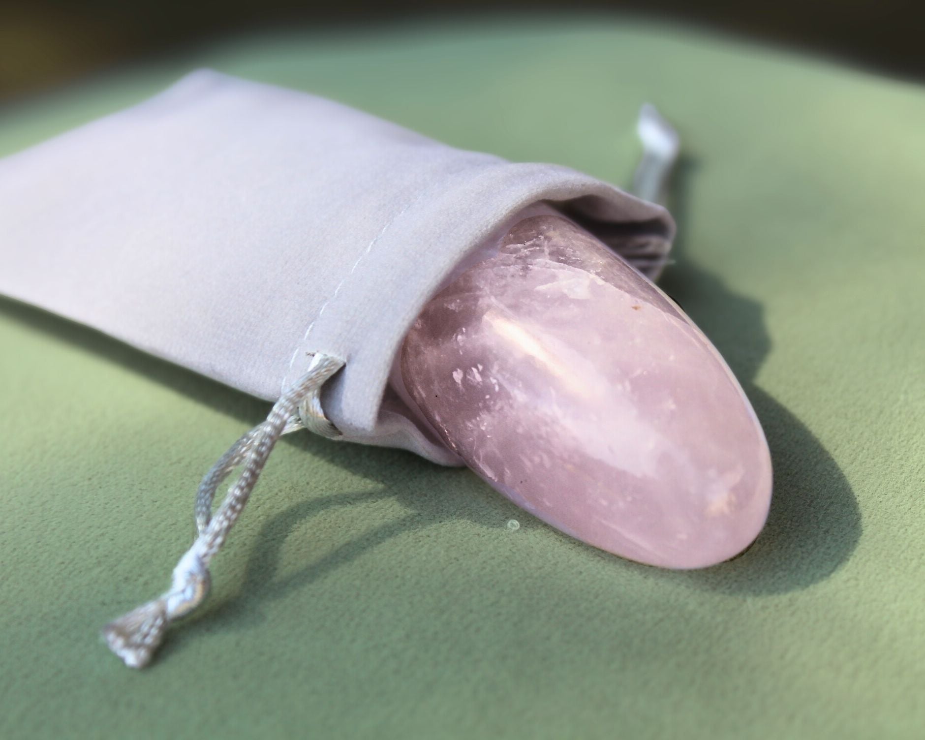 Rose Quartz palm stone partially out of a grey velvet drawstring bag, displayed on a sage green cushion — used for meditation, energy healing, and promoting love, compassion, and emotional balance.