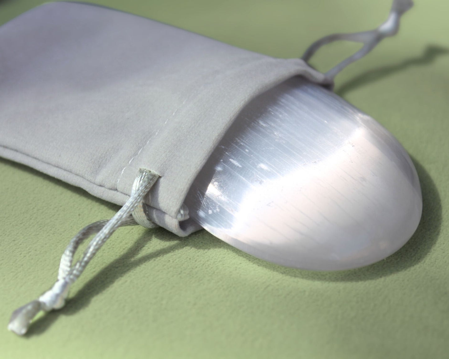 Selenite palm stone partially out of a grey velvet drawstring bag, displayed on a sage green cushion — used for meditation, energy cleansing, and promoting clarity, peace, and spiritual alignment.