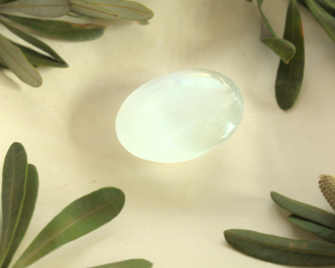 Selenite palm stone displayed on a light yellow background with natural greenery — used for meditation, energy cleansing, and promoting clarity, peace, and spiritual alignment.