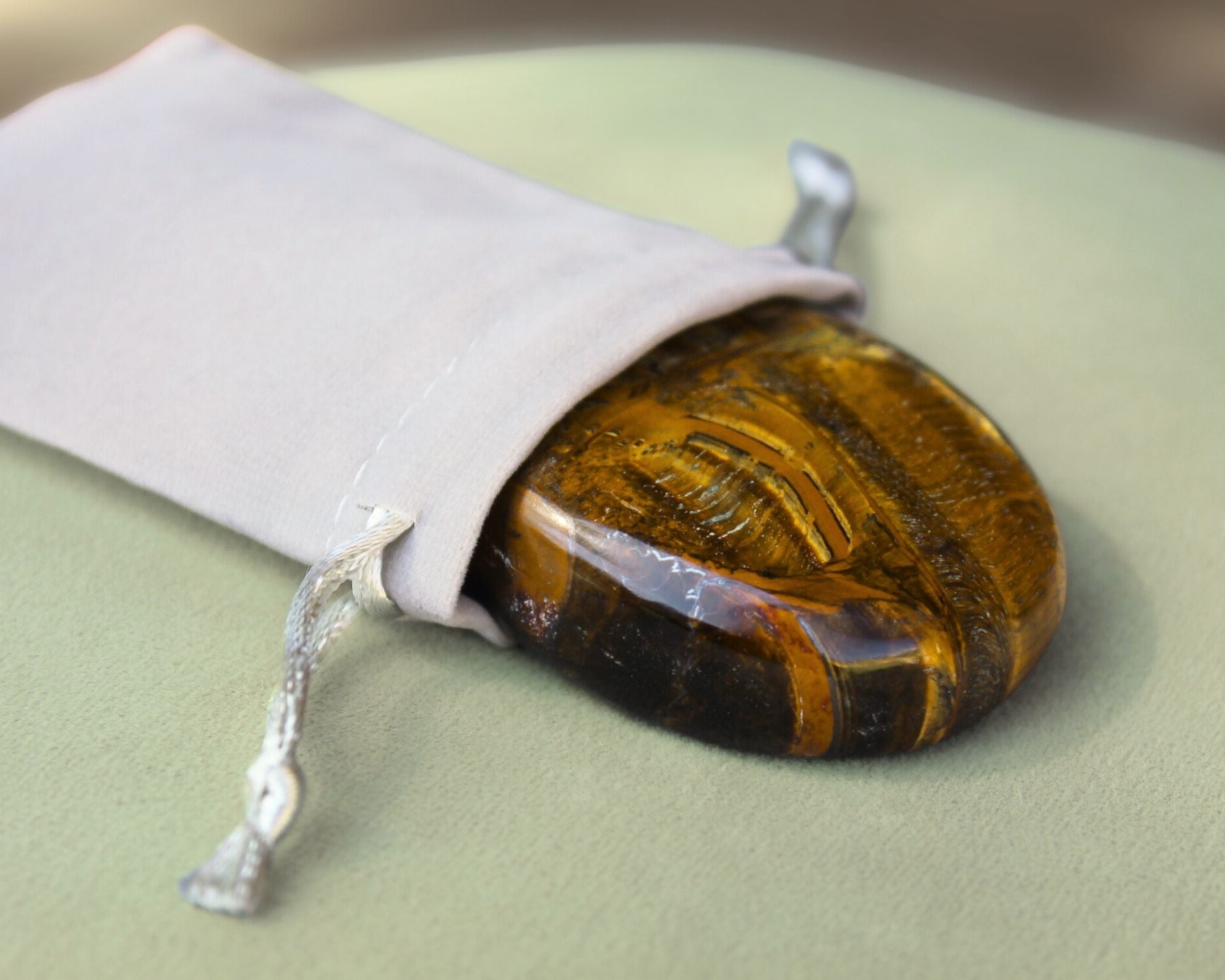 Tiger Eye palm stone partially out of a grey velvet drawstring bag, displayed on a sage green cushion — used for meditation, grounding, confidence, and courage.